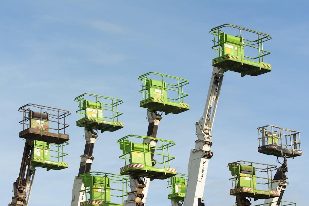A group of green cherry pickers extending against a clear blue sky in an industrial setting.