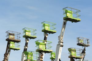 A group of green cherry pickers extending against a clear blue sky in an industrial setting.