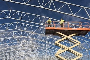 Two workers on a scissor lift at a construction site, framed by a metal structure against a blue sky.