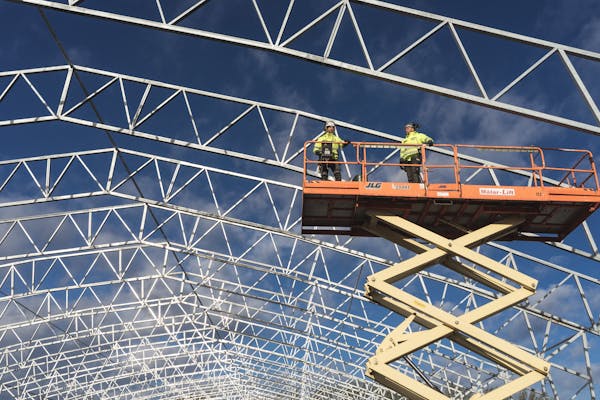 Two workers on a scissor lift at a construction site, framed by a metal structure against a blue sky.